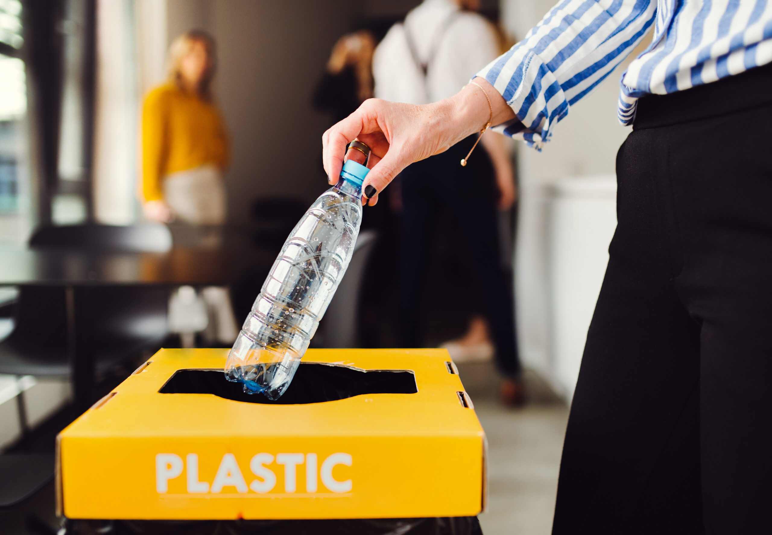 A female office worker throws an empty plastic bottle into a recycling bin labeled 'Plastic'